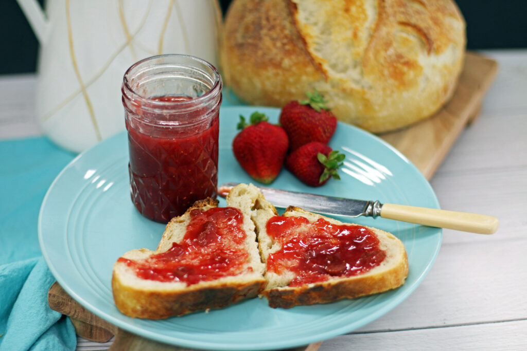 an open jar of strawberry jam with jam spread on two pieces of sour dough bread by an antique knife all set on a turquoise plate with a sourdough loaf of bread in the background