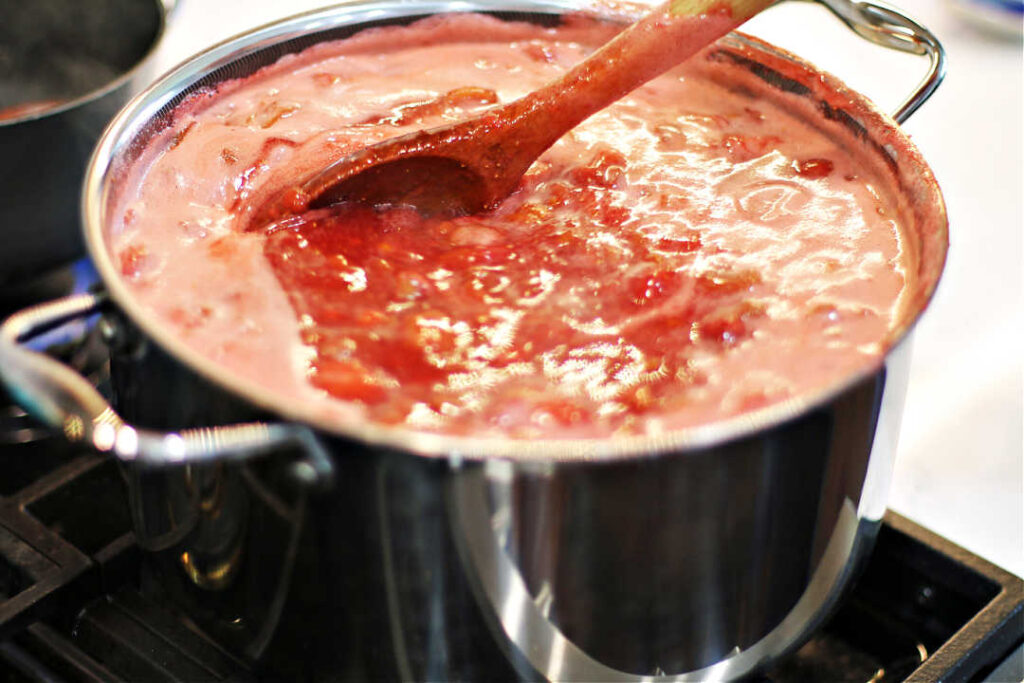 strawberry and rhubarb jam simmering in a large silver Dutch oven on a gas stove being stirred with a wooden spoon