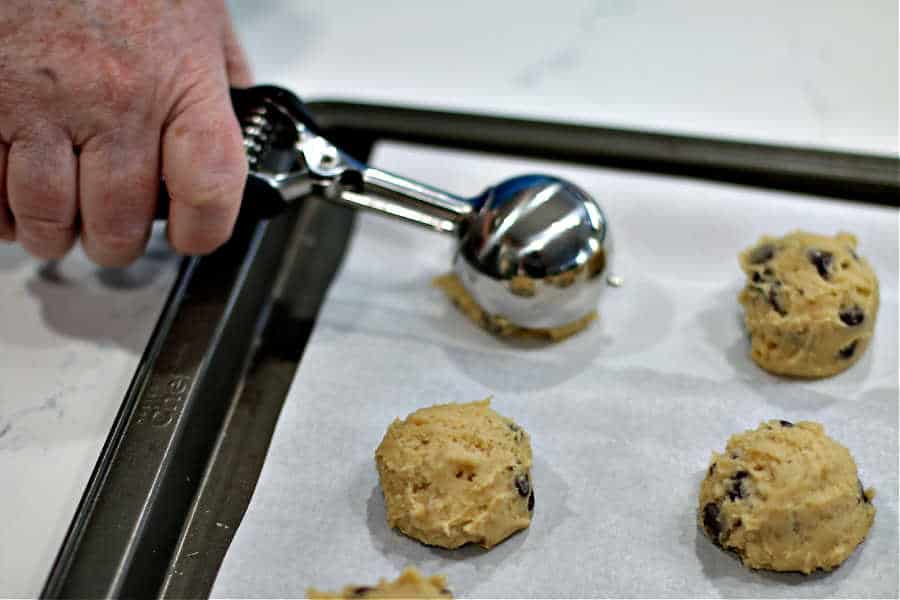 picture of a hand using a cookie scoop to place round mounds of dough onto a parchment paper covered cookie sheet 