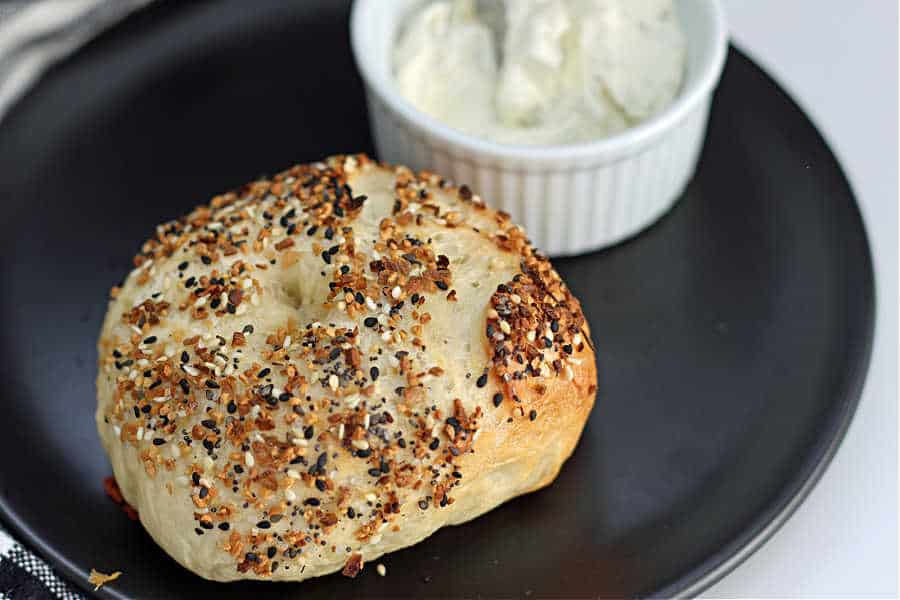 single sourdough bagel with Everything Bagel spice on top and a tub of sour cream on a black plate