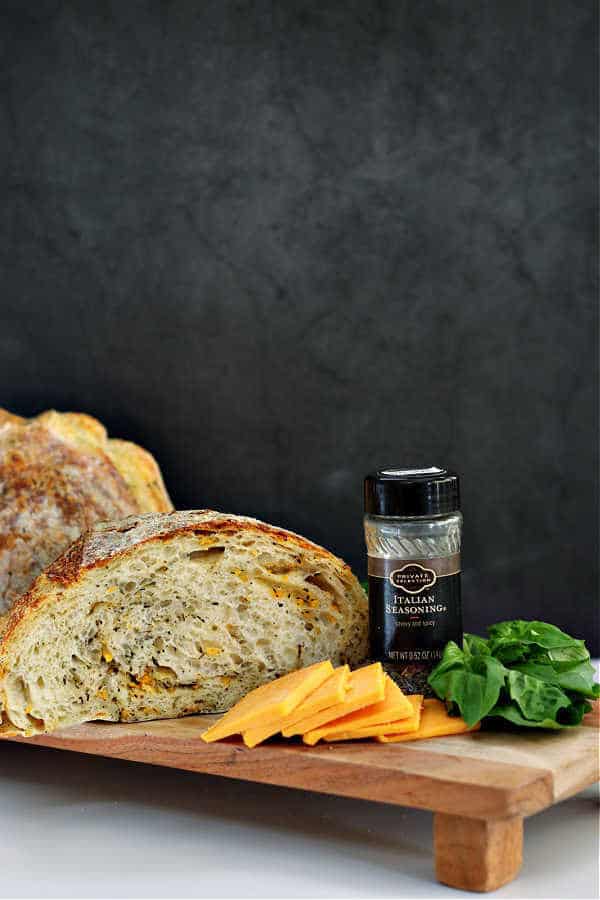 cut open italian herb and cheese sourdough artisan loaf on a wooden bread board with slices of cheddar cheese in front, fresh basil and a jar of italian seasoning off the the right side with a black background