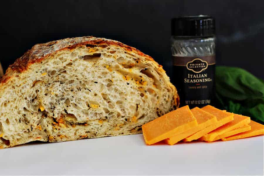 cut open italian herb and cheese sourdough artisan loaf with slices of cheddar cheese in front, fresh basil and a jar of italian seasoning off the the right side with a black background