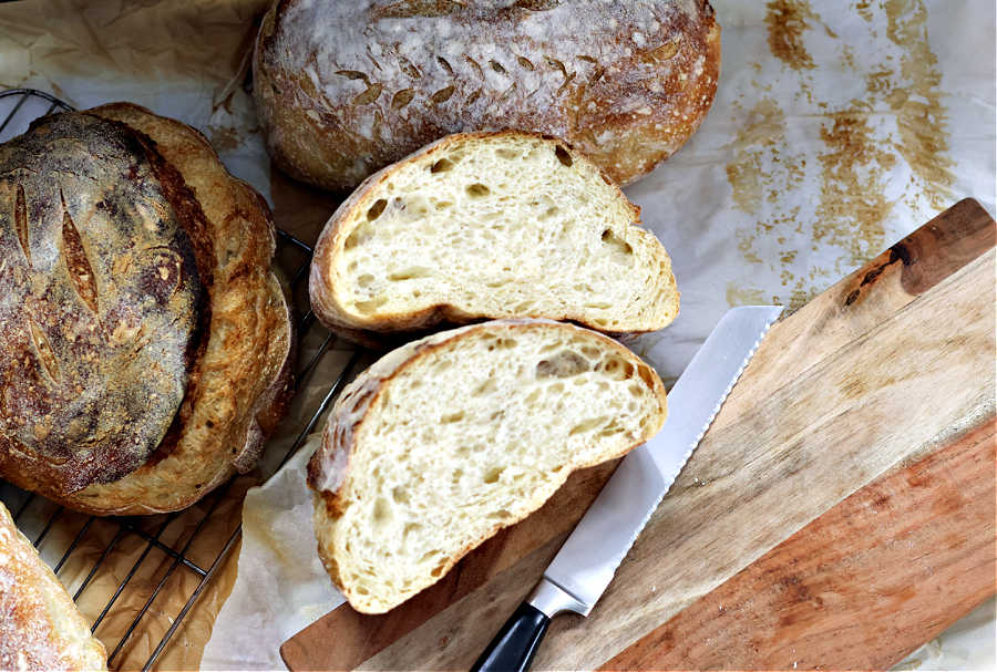 Sourdough boule cut in half showing large holes from high hydration surrounded by other breads, a brown wood cutting board and a silver knife with a black handle from the sour strawberry