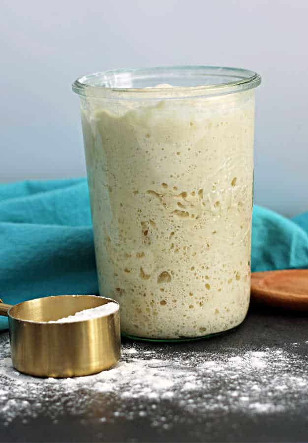 a full jar of fed active sourdough starter on a black counter with a brass measuring cup full of flower and a wooden spoon with a turquoise napkin in the background from the sour strawberry