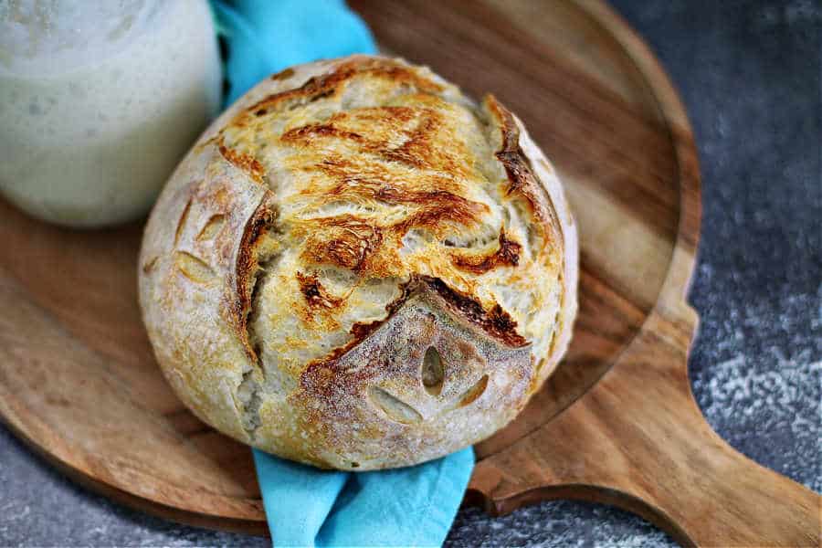 sourdough boule sitting on a round wood cutting board with a jar of starter beside it from the sour strawberry