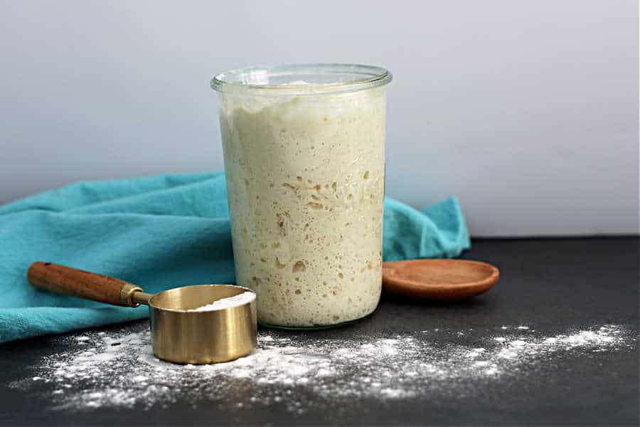 Picture of active sourdough starter in a jar with a measuring cup full of flour, a wooden spoon and a teal cloth napkin