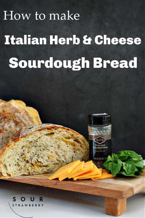 cut open italian herb and cheese sourdough artisan loaf on a wooden bread board with slices of cheddar cheese in front, fresh basil and a jar of italian seasoning off the the right side with a black background