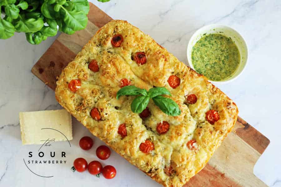 warm caprese sourdough focaccia bread with green pesto and cherry tomatoes, sliced on a wooden cutting board, with a large basil plant and a bowl of green pesto