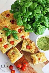 warm caprese sourdough focaccia bread with green pesto and cherry tomatoes, sliced on a wooden cutting board, with a large basil plant and a bowl of green pesto