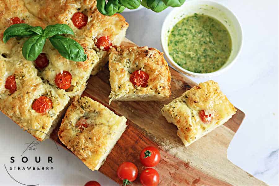 warm caprese sourdough focaccia bread with green pesto and cherry tomatoes, sliced on a wooden cutting board, with a large basil plant and a bowl of green pesto