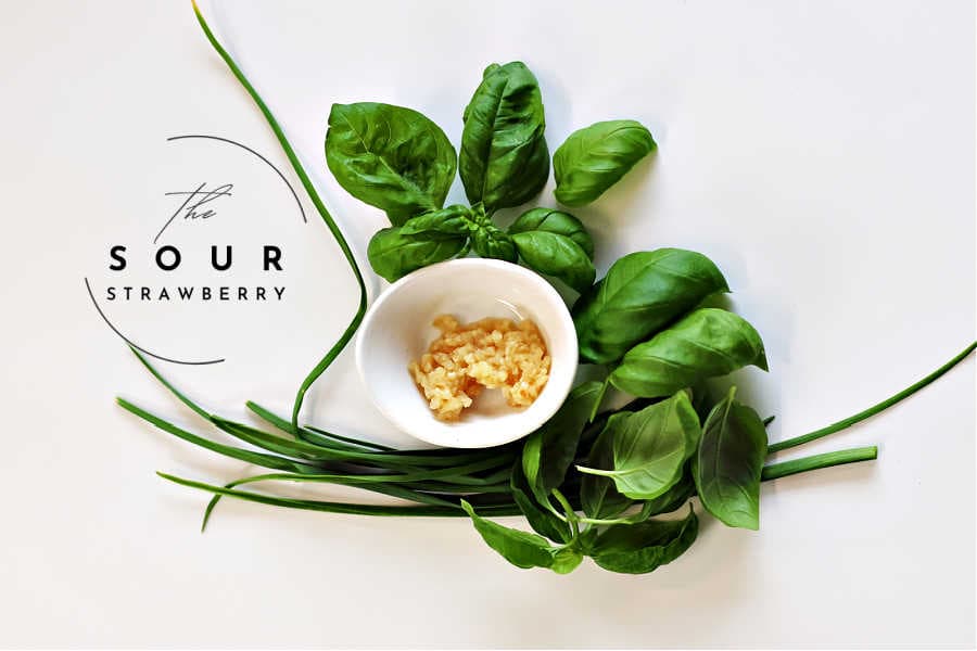Fresh garlic and basil ingredients for homemade pesto. A small white bowl of minced garlic is surrounded by fresh basil leaves and garlic scapes on a clean white background. Ideal for a quick and easy basil pesto recipe using fresh, aromatic ingredients.