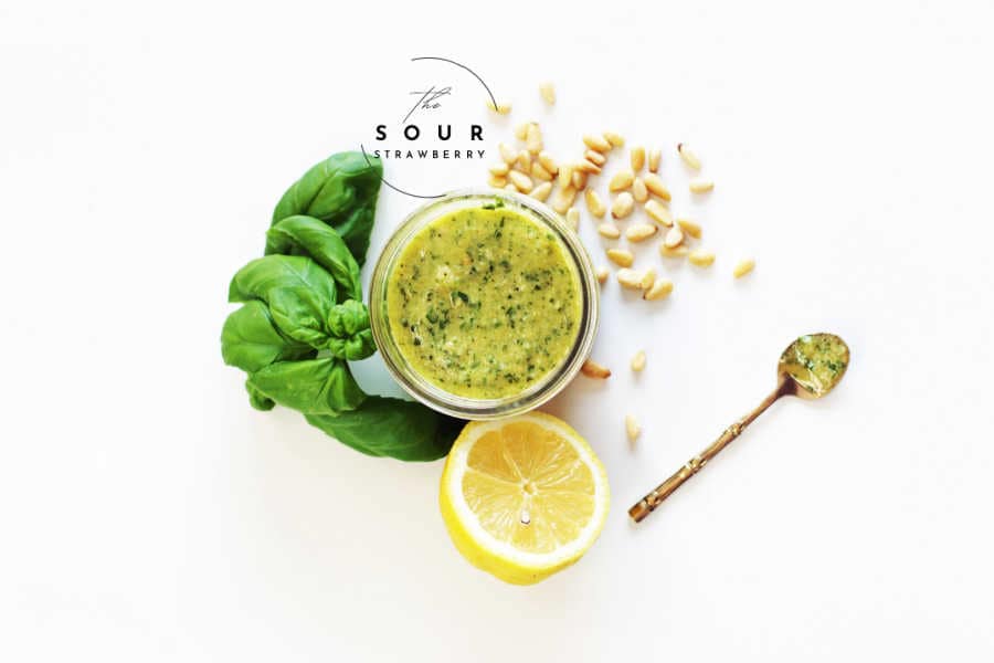 Jar of fresh homemade basil pesto surrounded by a bunch of fresh basil, a halved lemon, pine nuts, and a gold spoon with pesto on a white background. This vibrant green pesto sauce is perfect for pasta, sandwiches, or dipping, made with fresh basil, garlic, lemon juice, olive oil, and pine nuts—ideal for a quick and easy 5-minute pesto recipe.